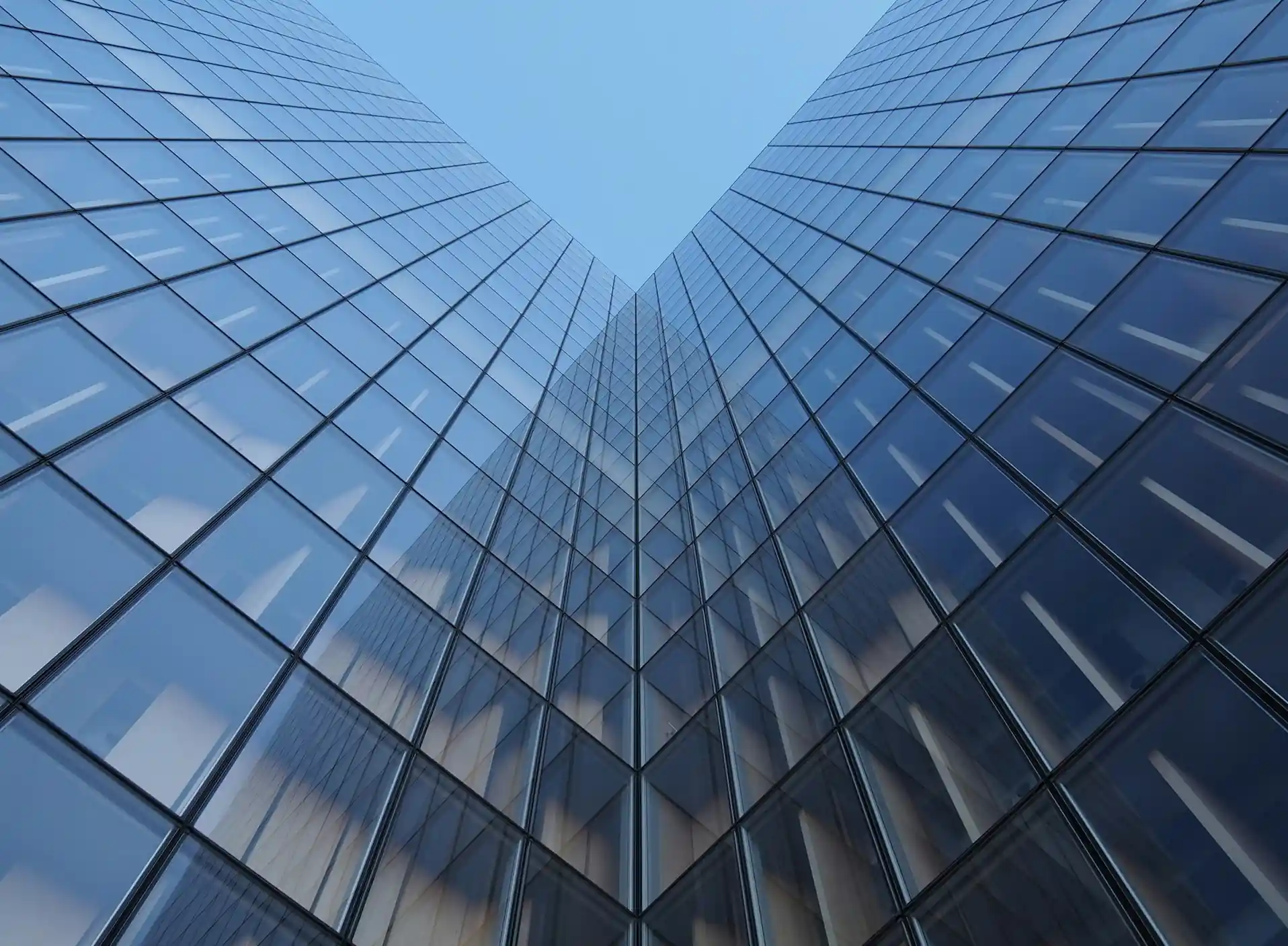Two glass towers seen from below, meeting against a pale blue sky